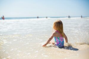 Young child in a colorful swimsuit sitting in shallow ocean water at the beach.