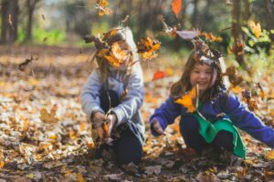 Two young children playing in a pile of autumn leaves in a sunlit wooded area.